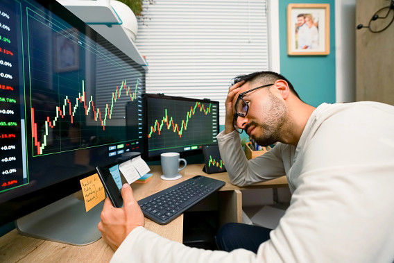 A man sitting at a desk, looking stressed while examining multiple computer screens displaying stock market charts with fluctuating green and red lines. He holds a smartphone in one hand and runs his other hand through his hair, indicating concern.