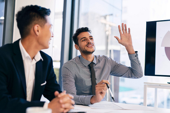 Two business professionals in conference room, one raising hand during meeting with pie chart presentation on screen in background.