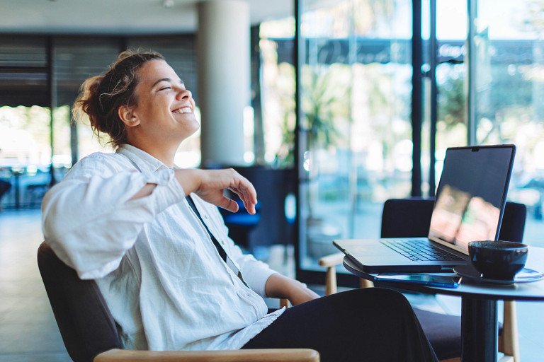 Person in white shirt working on laptop at a table with a coffee cup, seated beside large windows with a view bright outdoor scenery