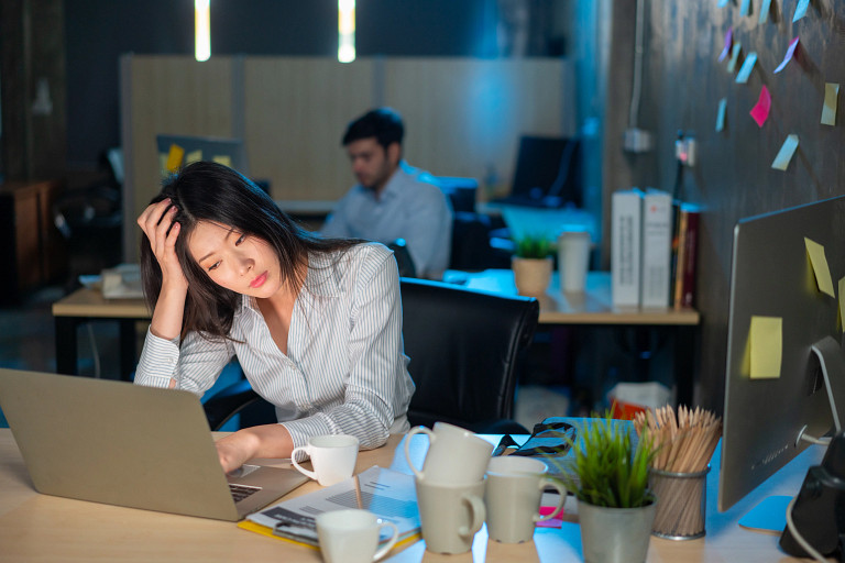 Modern office workspace with two people working at desks; foreground person focused on laptop with hand on head, surrounded by coffee mugs, papers, and a plant; background includes another worker and a wall covered in sticky notes
