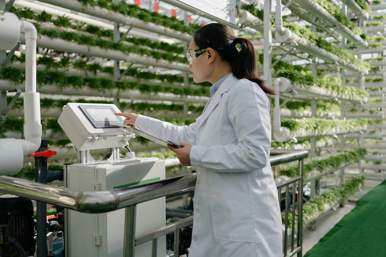 A scientist in a white lab coat and safety goggles operates a touchscreen device in a hydroponic farm. Rows of green plants grow in vertical pipes in the background, showcasing a high-tech agricultural environment.