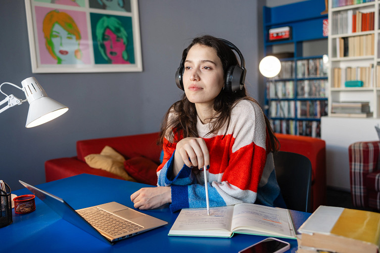 A young woman wearing headphones sits at a blue desk with an open notebook, pen in hand, and a laptop in front of her. She appears focused and thoughtful. The background features colorful wall art, shelves with books, and a red sofa.