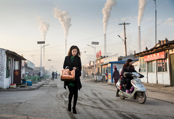 A woman walks through a village street with industrial smokestacks emitting smoke in the background. The scene highlights the contrast between daily life and the impact of industrial pollution. Other people are visible in the background, including someone riding a scooter. The setting features small shops and buildings, emphasizing a rural or semi-urban environment under the shadow of industrial activity.