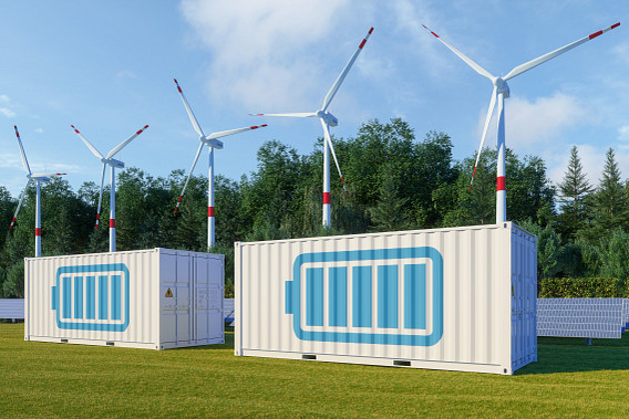 A renewable energy setup featuring wind turbines, solar panels, and two white battery storage containers with blue battery icons. The scene is set in a grassy area with trees in the background and a clear blue sky above.