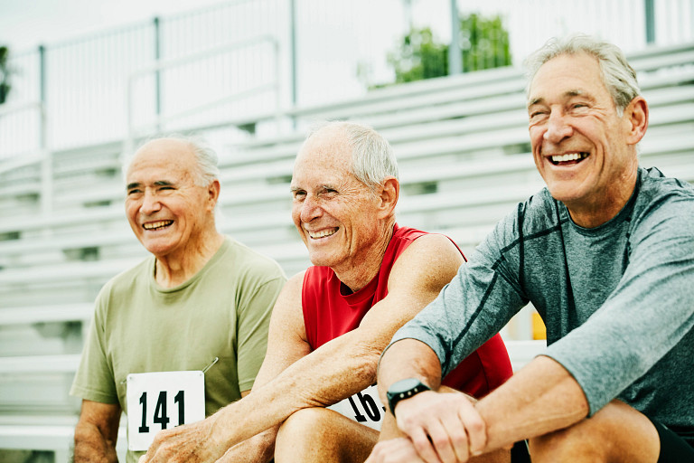 Three senior men sitting on bleachers, smiling and enjoying themselves. Two are wearing numbered bibs, suggesting participation in a sporting event, while one is dressed in casual athletic wear. The scene is outdoors with a bright and cheerful atmosphere, emphasizing active and healthy aging.