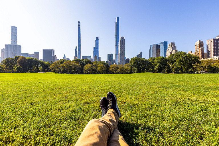 A person is lying on a grassy field in a park, with their legs and black shoes visible in the foreground. The scene overlooks a city skyline with tall skyscrapers under a clear blue sky. Trees border the grassy area, adding a natural contrast to the urban background. The perspective creates a relaxing and peaceful atmosphere.