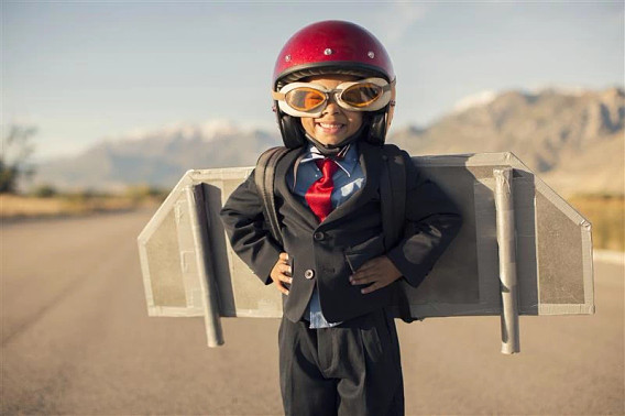 This image depicts a young child wearing a business suit, tie, and a red motorcycle helmet. The child is standing in a desert-like landscape, holding what appears to be a small cardboard box or structure in front of them. The image conveys a sense of whimsy and imagination, with the child playing at being a professional or businessman in an unexpected outdoor setting.