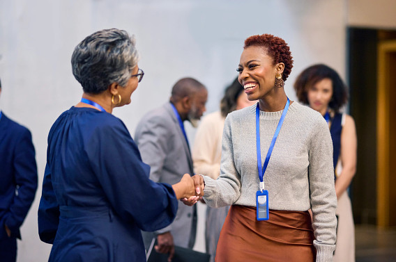 Two women are shaking hands and smiling at each other in a professional setting. Both wear name tags, indicating a networking or business event. In the blurred background, other people are engaged in conversations. The scene conveys a positive and friendly interaction.